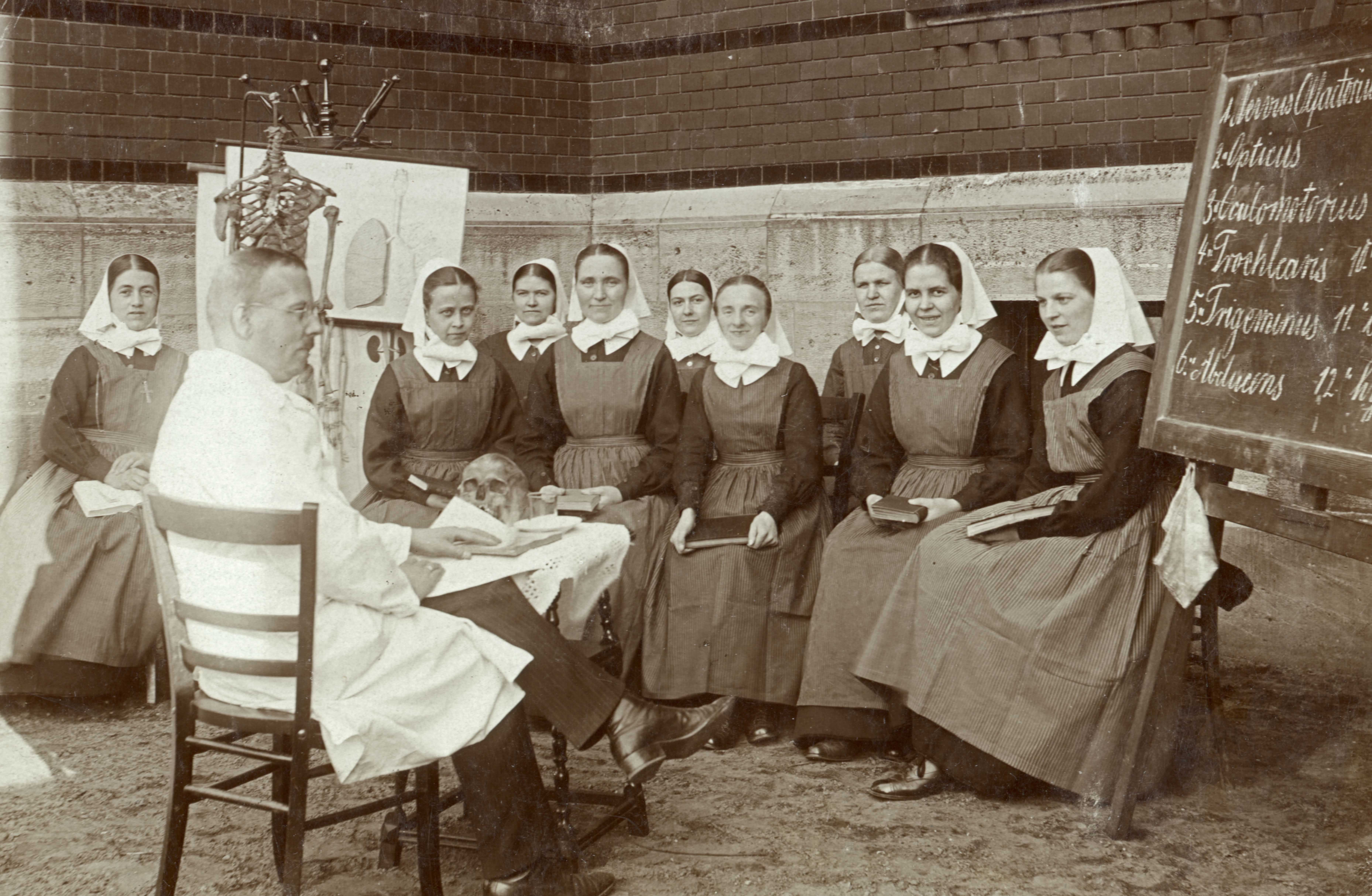 (Schwarz-Weiss-Foto vpn 1926) Arzt im Kittel unterichtet eine Gruppe von Diakonissen. Rechts im Bild steh eine Tafel, links eine Abbildung von der Lunge sowie ein Skelett