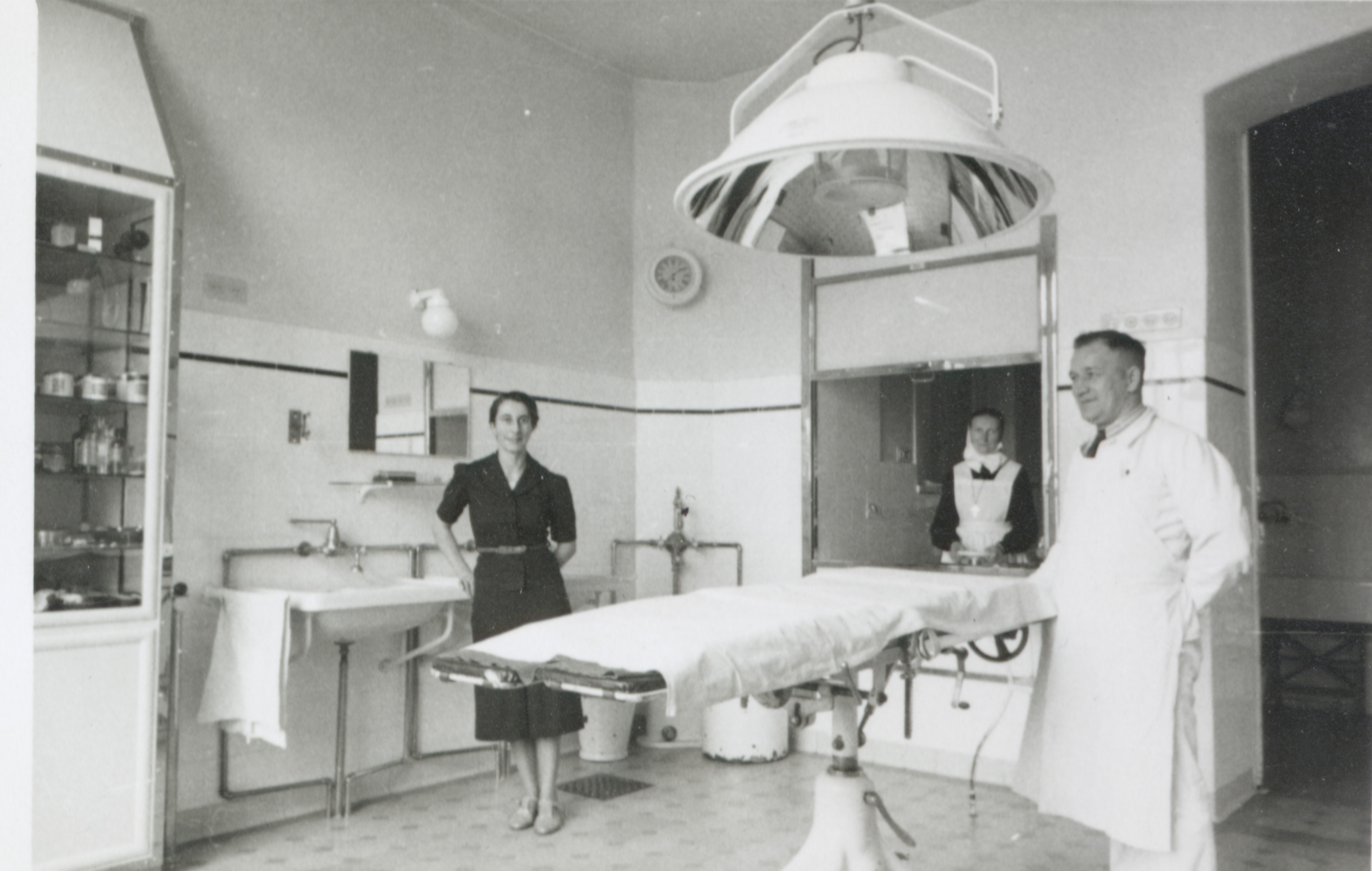 Ein Arzt und eine Frau im dunklen Klein stehen in einem Op-Saal. Eine Diakonisse schaut hinter einer Scheibe in den Raum. Alle Lachen.  (Schwarz-Weiss-Foto)
