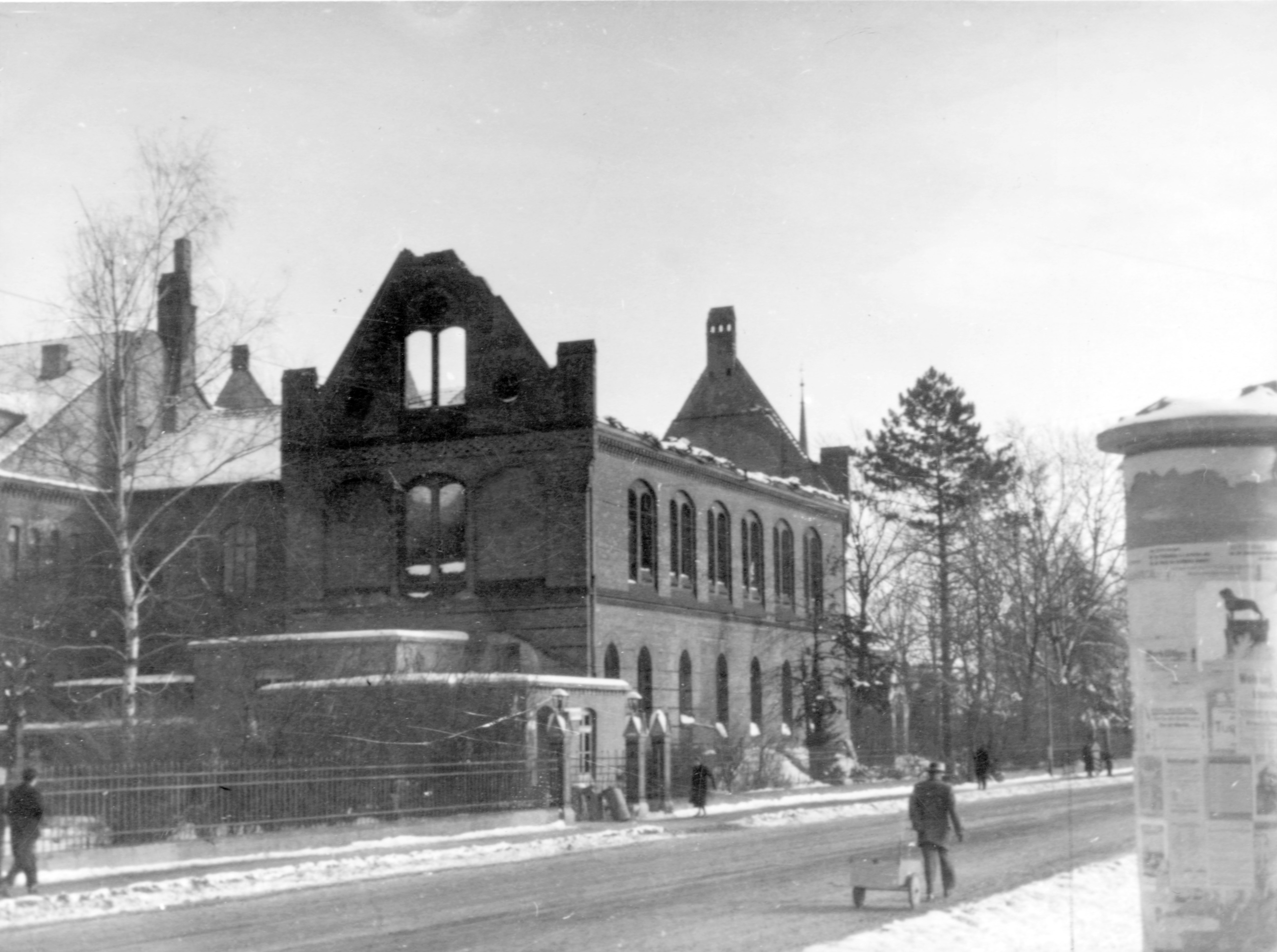 Bild vom Krankenaus mit der Stiftkapelle 1944. Es liegt Schnee, das Dach des Huases fehlt, ein Mann läuft mit einem Handwagen die Strasse entlang, rechts ist eine Litfasssäule zu sehen.(Schwarz-Weiss-Foto)