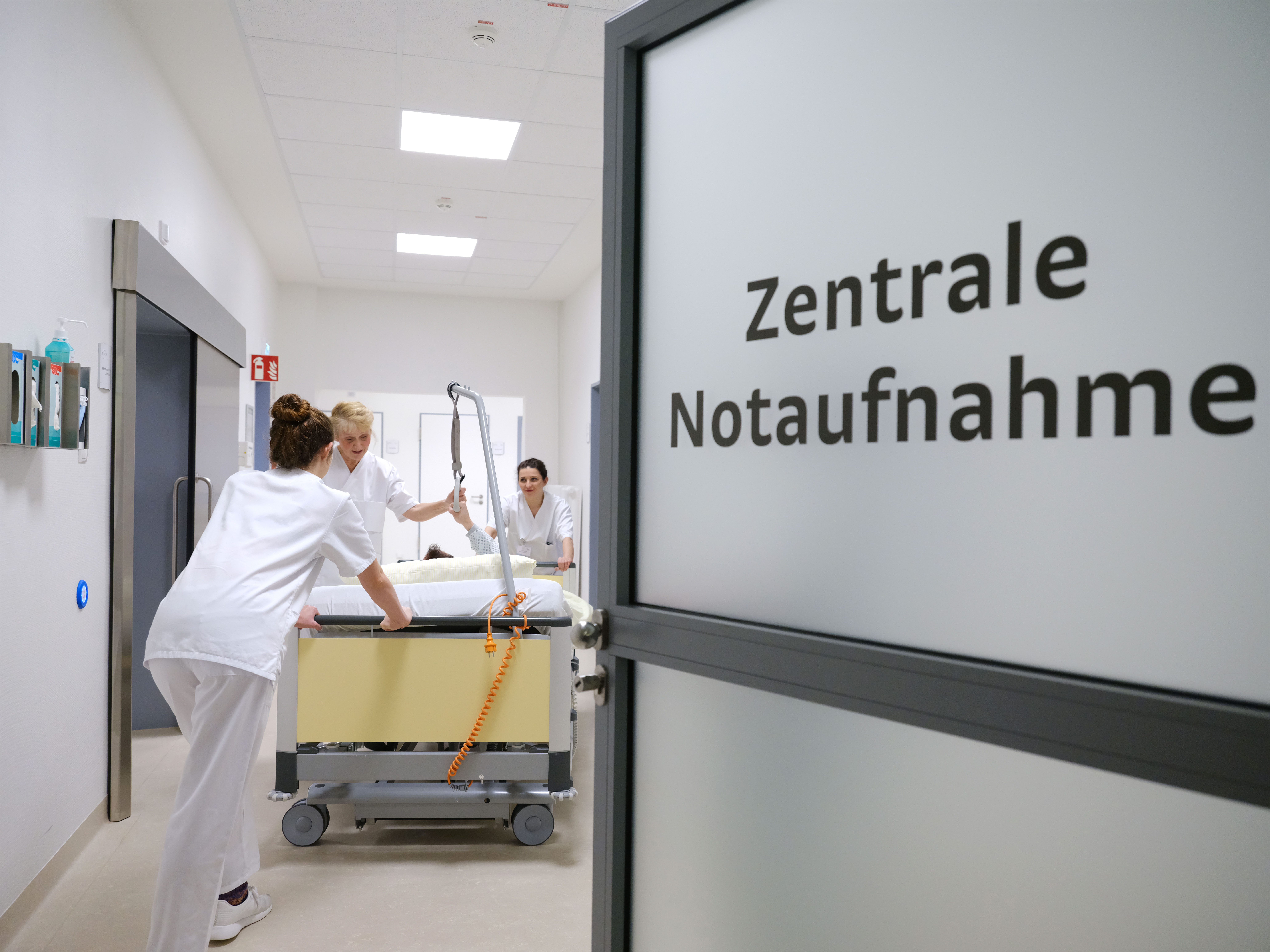 Eine halb geöffnete Tür mit Aufschrift Zentrale Notaufnahme aus Milchgrlas. Ein Bett wird vom Pflegepersonal  in den Raum geschoben