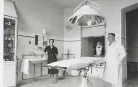 Ein Arzt und eine Frau im dunklen Klein stehen in einem Op-Saal. Eine Diakonisse schaut hinter einer Scheibe in den Raum. Alle Lachen.  (Schwarz-Weiss-Foto)