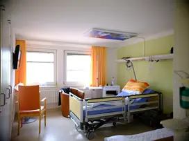 Ein Zimmer der Palliativstation, die wand ist hell grün, die Vorhänge organe, es herrscht eine freundliche und ruhige Atmosphäse