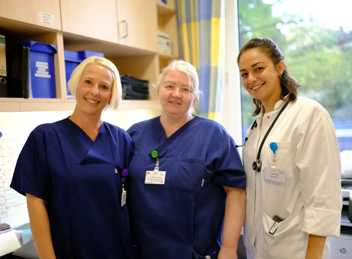 zwei Krankenpflgerinnen und eine Assistenzärztin stehen zusammen und schauen lachend in die Kamera.