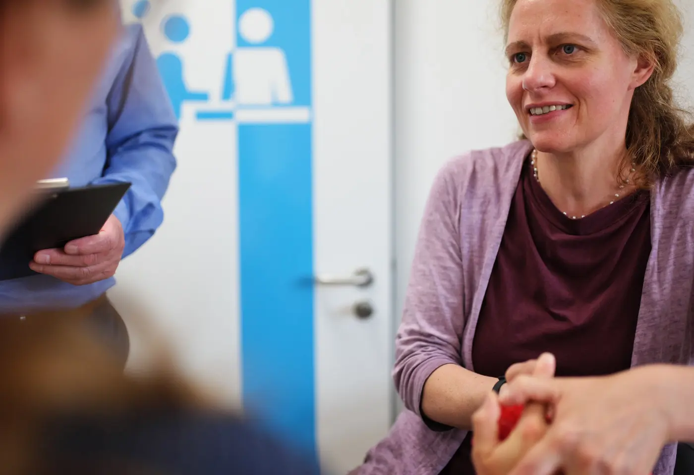 Weibliche Fachkraft mit blondem Haar und lila Strickjacke spricht mit einem Patient, dieser ist von hinten zu sehen