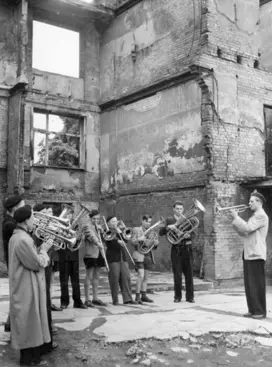 Eine Blaskapelle aus 10 Jungen und Männern spielt auf verschiednenen Blasinstrumenten vor einem stark zerbomten Gebäude (Schwarz-Weiss-Foto)
