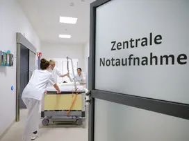 Eine halb geöffnete Tür mit Aufschrift Zentrale Notaufnahme aus Milchgrlas. Ein Bett wird vom Pflegepersonal  in den Raum geschoben